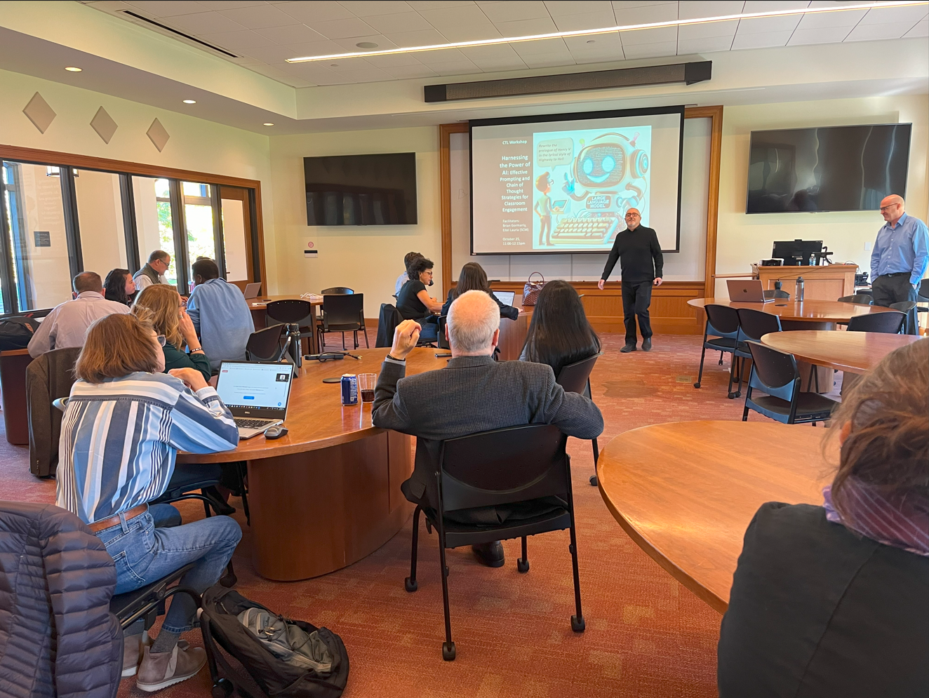 A classroom workshop with participants seated at round tables, listening to two presenters. A slide titled