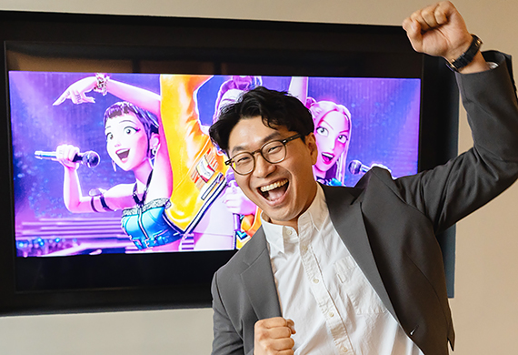 Dr. Sang-Keun Yoo, who teaches with K-Pop, Film, and Dramas this fall, poses for a portrait in front of a TV showing the Kpop Demon Hunters movie.