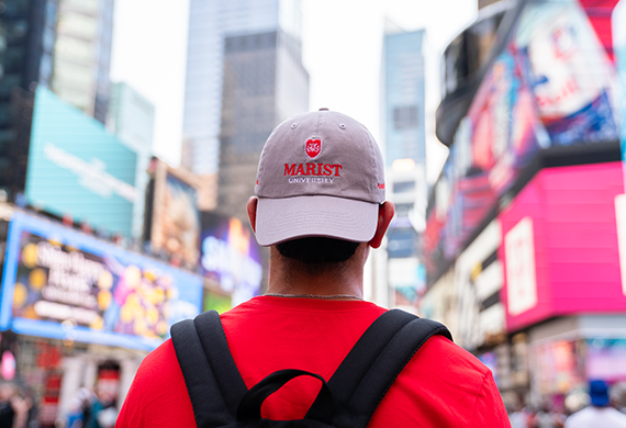 Image of the back of a student standing in Time Square wearing a hat that says 