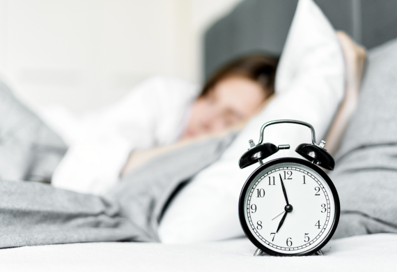 Image of a person sleeping in bed with an alarm clock in the foreground. 